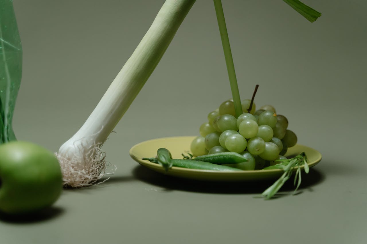 A minimalist arrangement of green grapes, leeks, and an apple on a plate.
